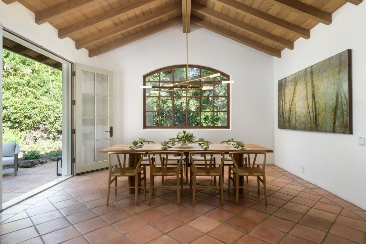 A dining room with an exposed beam ceiling, a window, a painting, and a dining table surrounded by chairs.