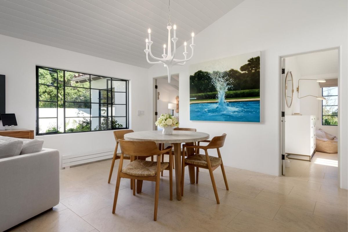 A breakfast nook with a painting, a window, a chandelier, and a round table surrounded by chairs.