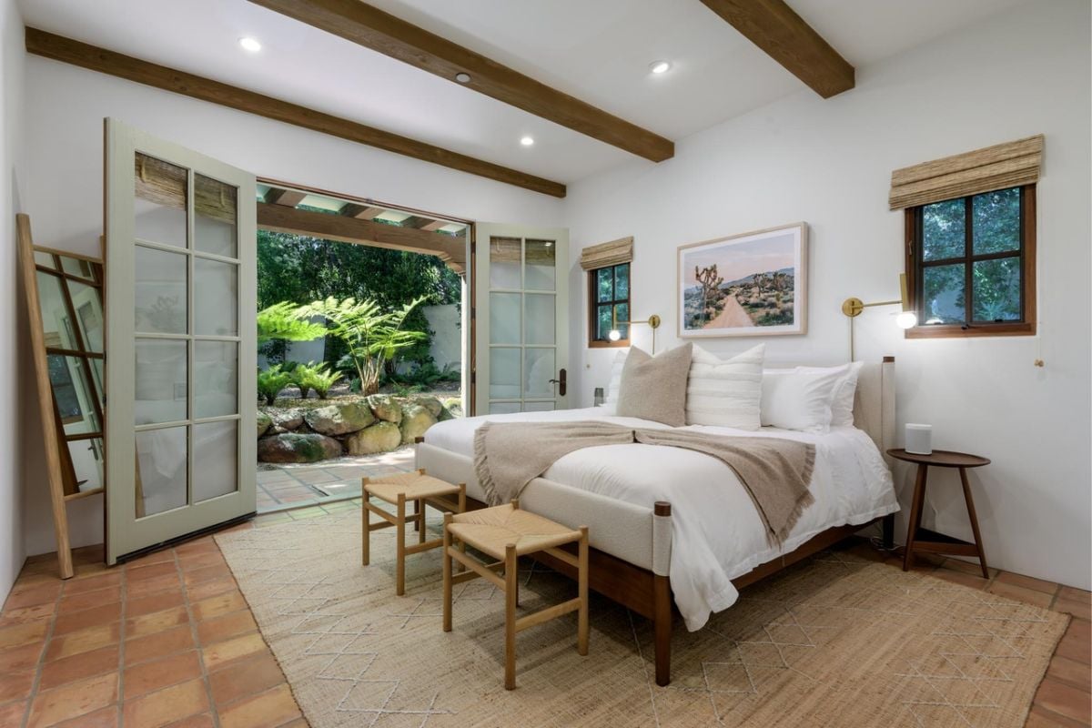 A bedroom with a bed, chairs, a side table, a painting, windows, and a beamed ceiling.
