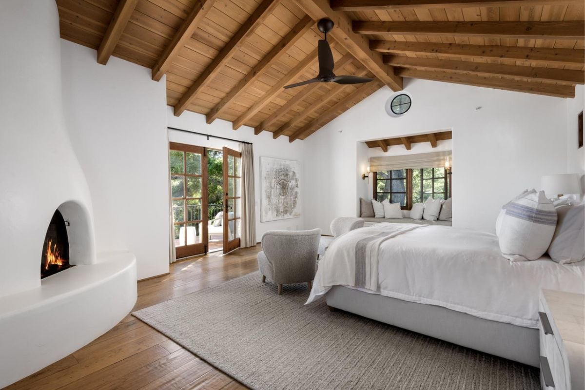 A bedroom with a bed, an exposed beam ceiling, a fireplace, a couch, a window, and window seating.