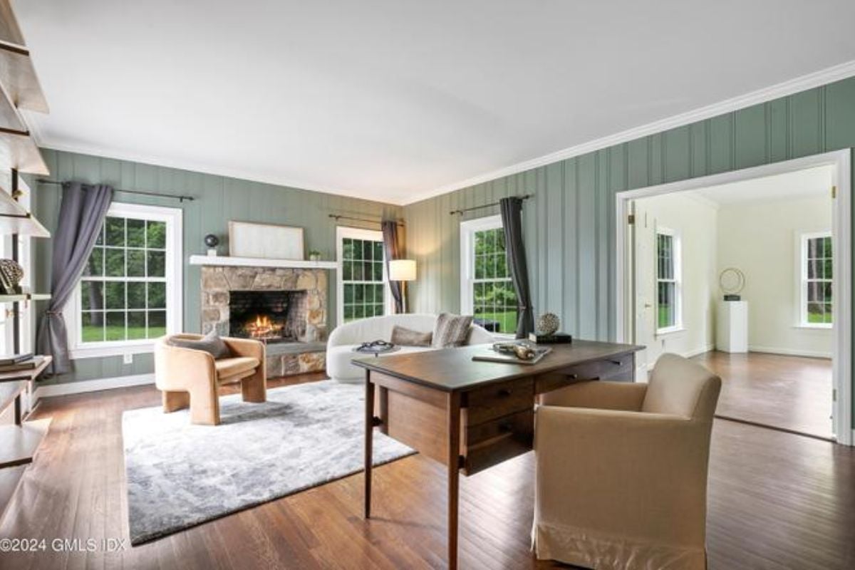 A home office with a rug, windows, fireplace, couch, shelves, painting, desk, and chair.