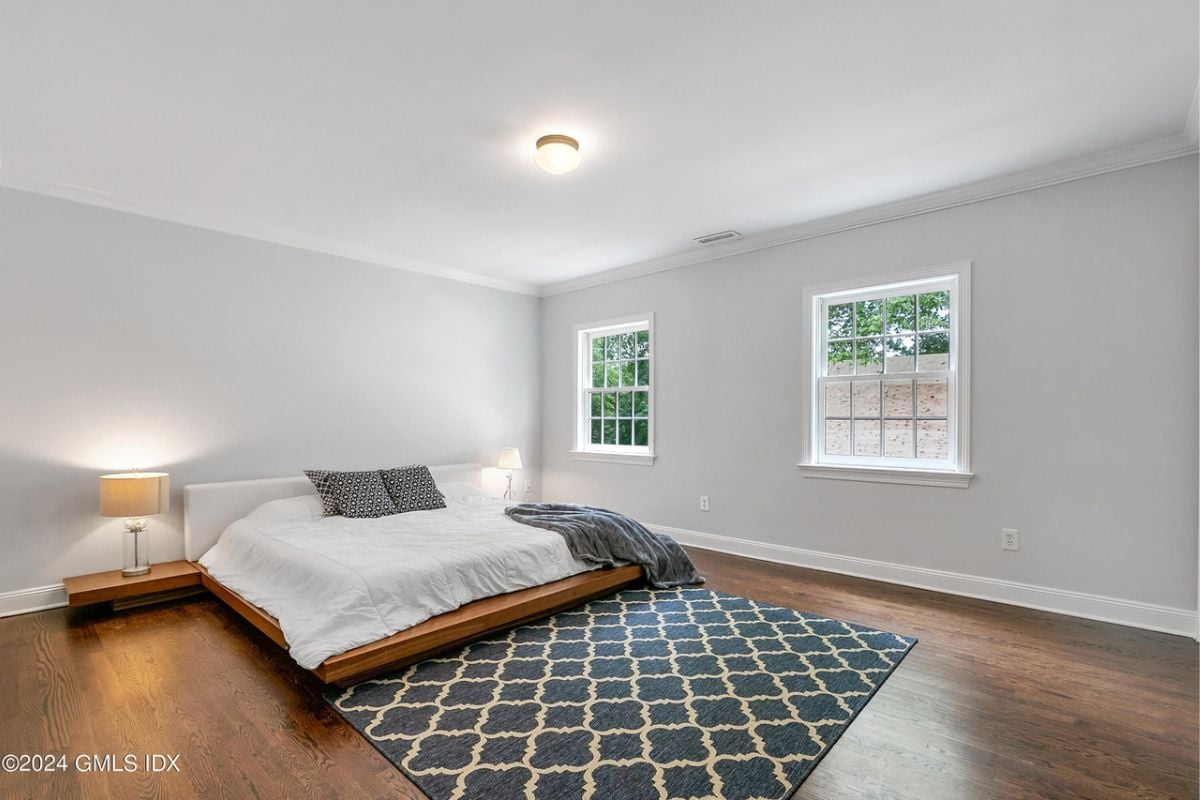 A bedroom with a bed, windows, white ceiling, white walls, a rug, and a wooden floor.
