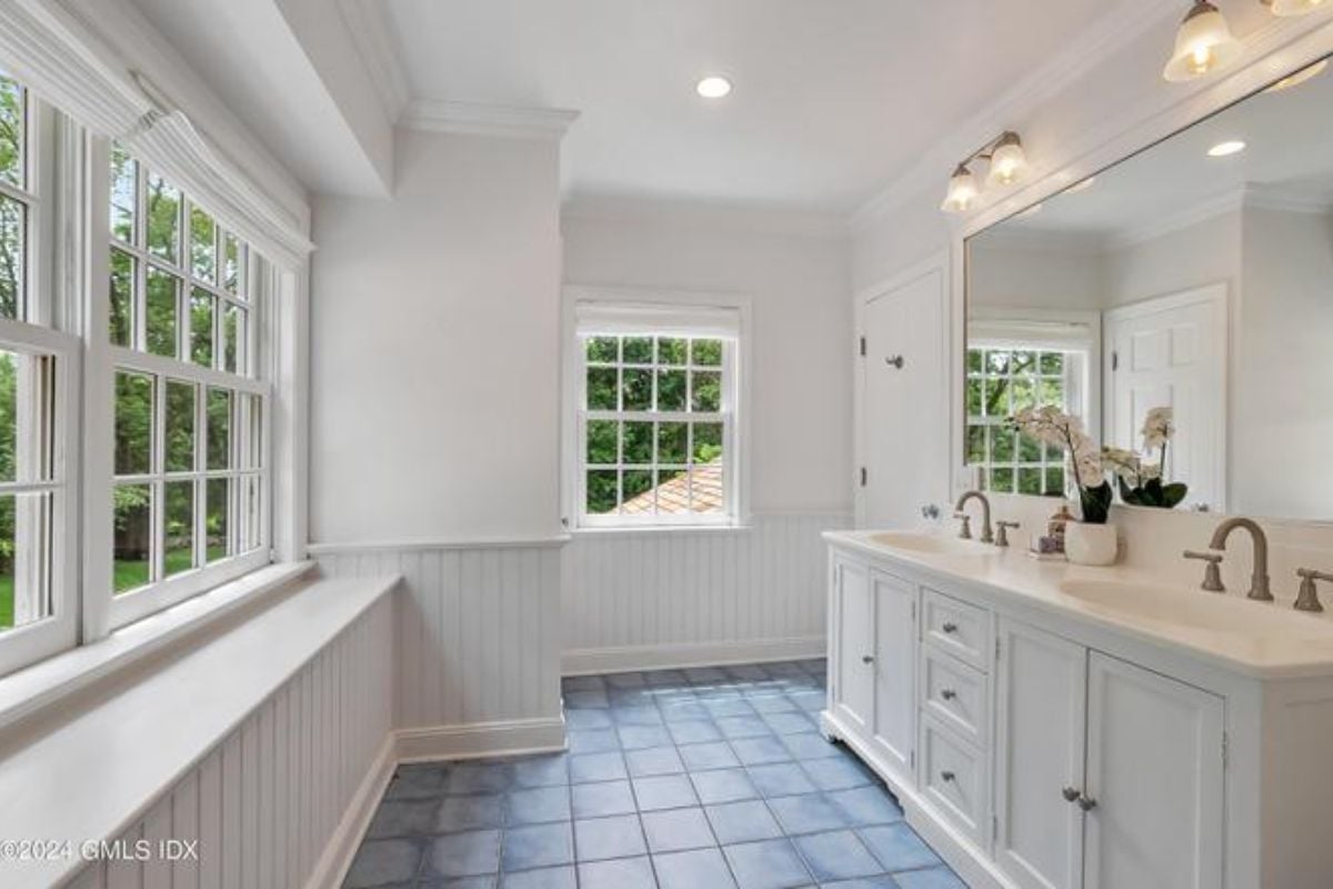 A bathroom with a tiled floor, countertop, sink, mirror, cabinets, and windows.