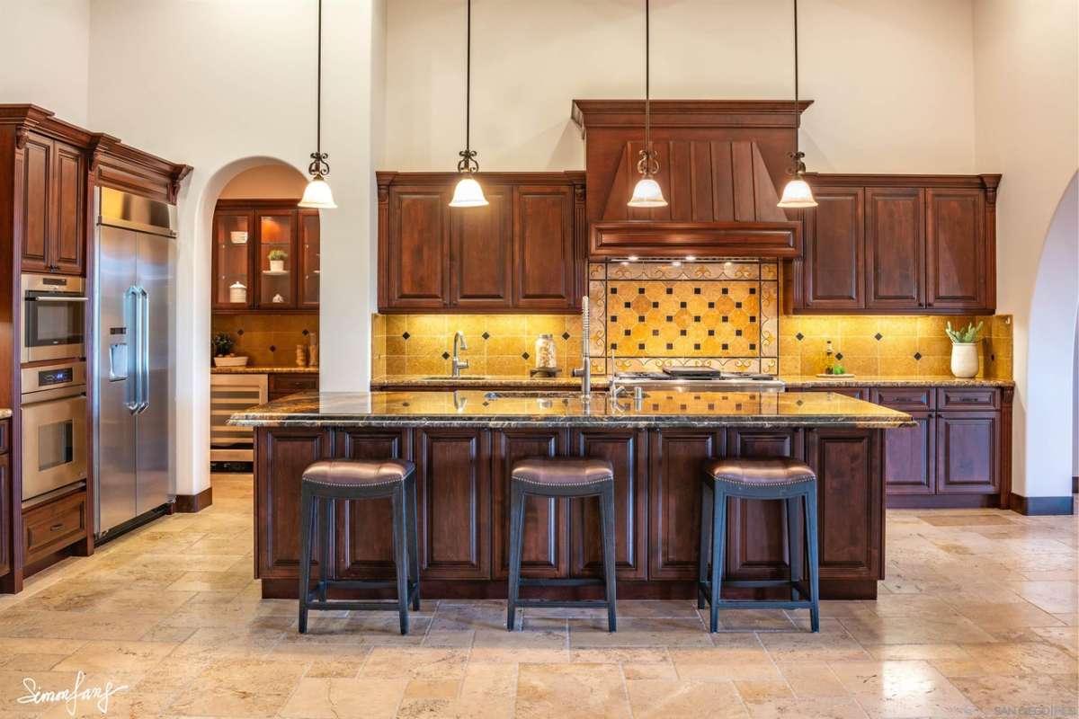 A kitchen with a tiled floor, countertop, chairs, sink, wooden cabinets, and kitchen appliances.