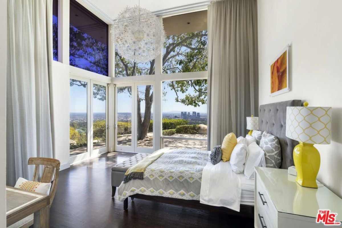A bedroom with a bed, nightstand, chandelier, windows, painting, desk, and chair.