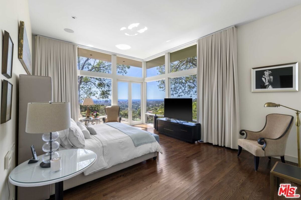 A bedroom with a bed, hardwood floor, windows, side table, chairs, and TV.