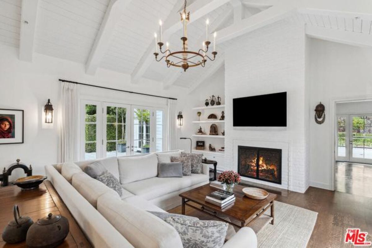A living room with a fireplace, TV, white couch, center table, chandelier, and exposed beam ceiling.