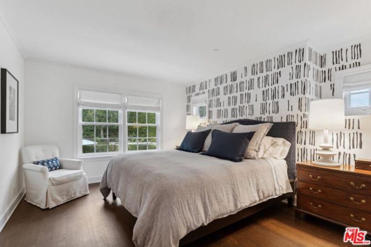 A bedroom with a bed, wallpaper, cabinets, windows, a lampshade, and a chair.