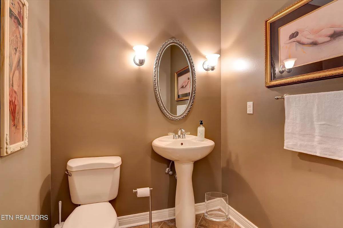 Powder room with a pedestal sink and brown walls graced with an oval mirror and framed artworks.