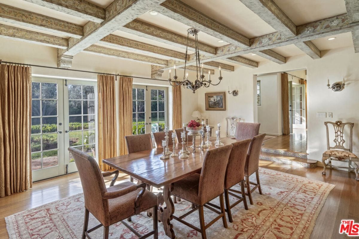 A dining room with an exposed beam ceiling, chandelier, windows, rug, and a dining table surrounded by chairs.