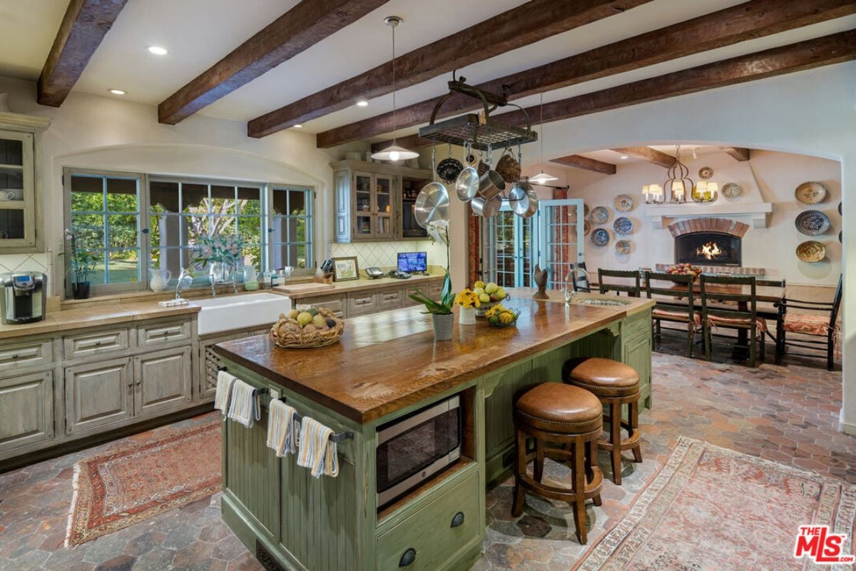 A dining area and kitchen with an exposed beam ceiling, fireplace, dining table, chairs, bench, island, countertop, sink, and cabinets.