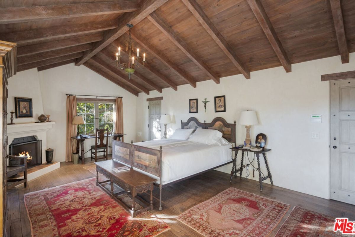 A bedroom with an exposed beam ceiling, bed, side table, fireplace, desk, chair, and window.