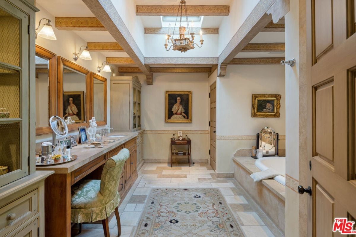 A bathroom with a countertop, powder area, chair, cabinets, paintings, chandelier, bathtub, and exposed beam ceiling.