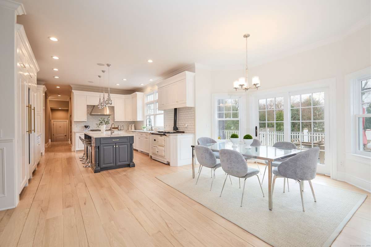 A kitchen with wooden floors, a rug, island, countertop, white cabinets, windows, and a dining table surrounded by chairs.