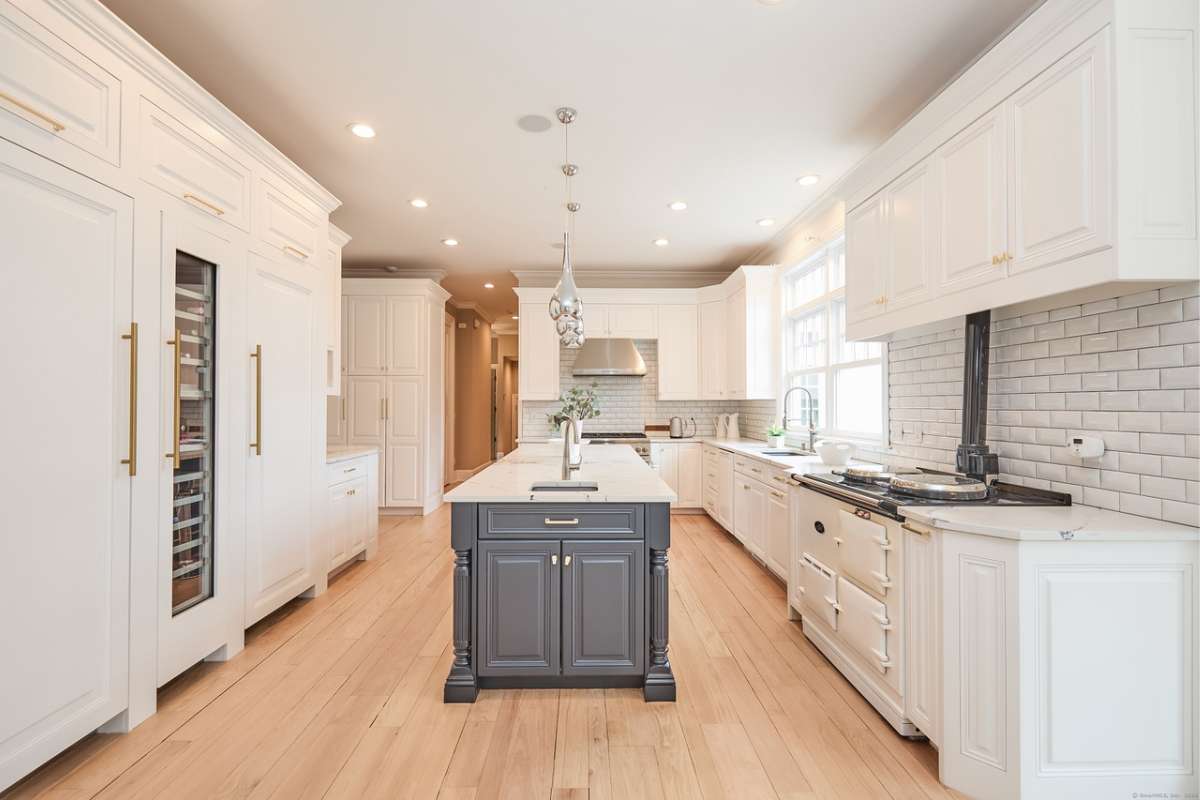 A kitchen with an island, countertop, sink, white cabinets, windows, and kitchen appliances.