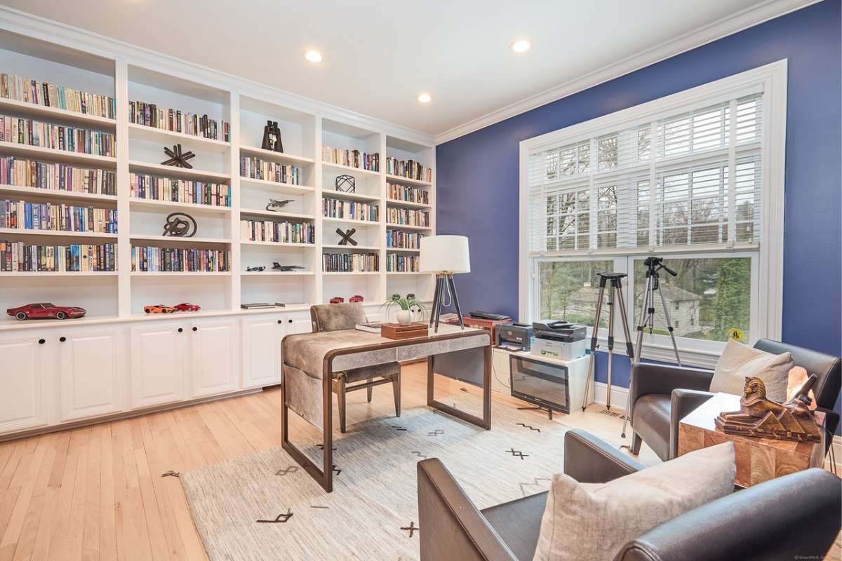 A home office with a wooden floor, desk, chairs, bookshelf, windows, and a white ceiling.