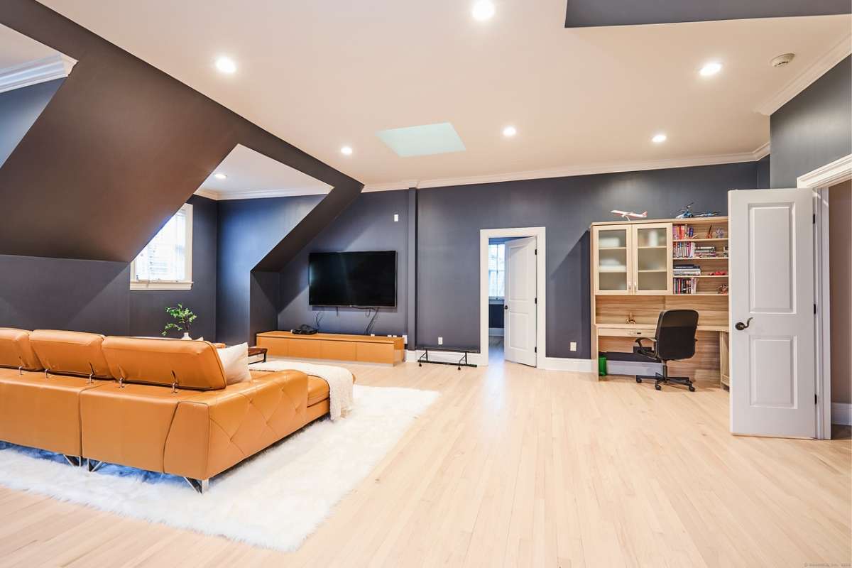 A lounge with a brown couch, TV, wooden floor, chair, shelf, and window.