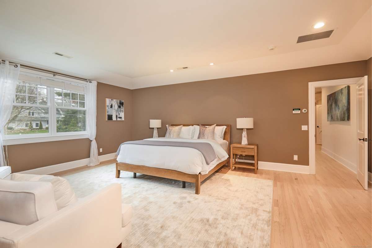 A bedroom with a bed, rug, wooden floor, nightstand, lampshade, windows, couch, and a white ceiling.