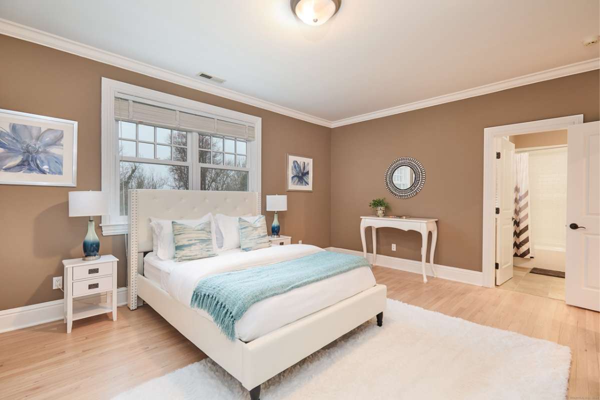 A bedroom with a bed, wooden floor, rug, nightstand, lampshade, windows, and a white ceiling.