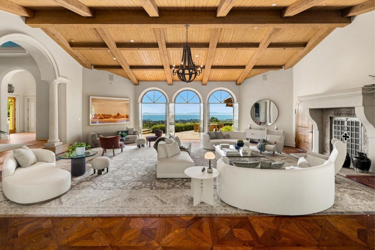 A living room with an exposed beam ceiling, windows, white couches, a chandelier, chairs, and coffee tables.