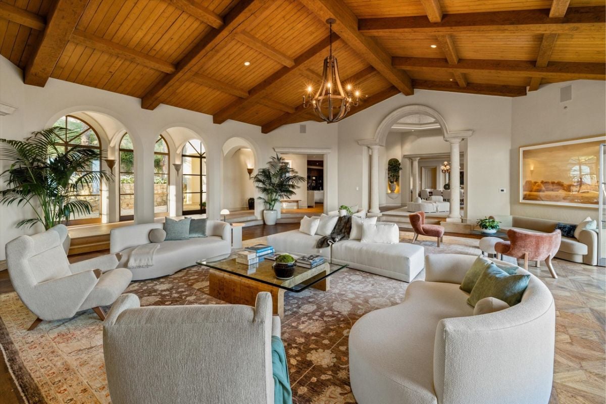A living room with an exposed beam ceiling, windows, white couches, a chandelier, chairs, and coffee tables.