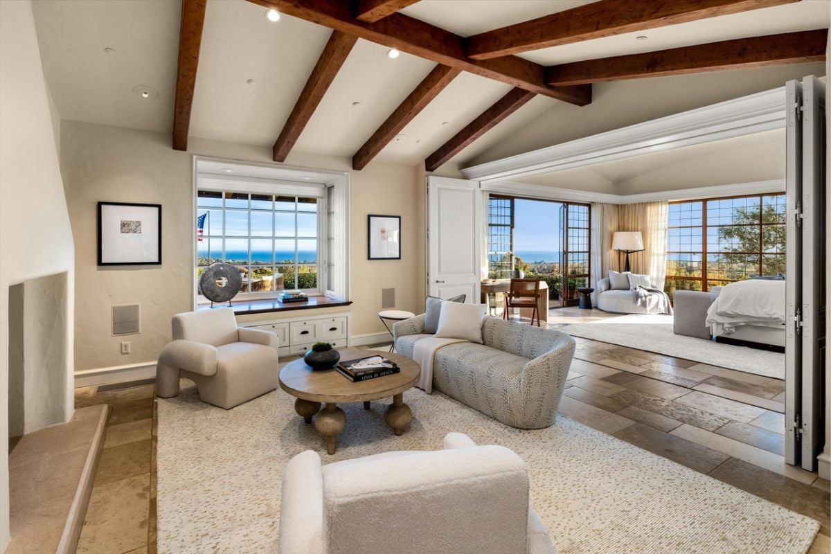 A living room with an exposed beam ceiling, fireplace, rug, windows, couches, and a coffee table.