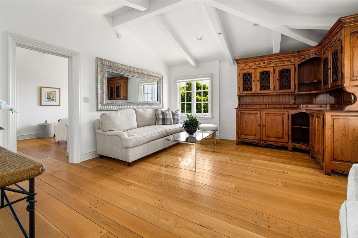 A living room with an exposed beam ceiling, glass coffee table, wooden floor, and cabinets.