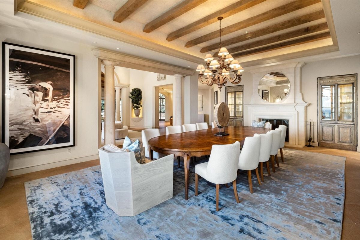 A dining room with an exposed beam ceiling, painting, rug, chandelier, and dining table surrounded by chairs.