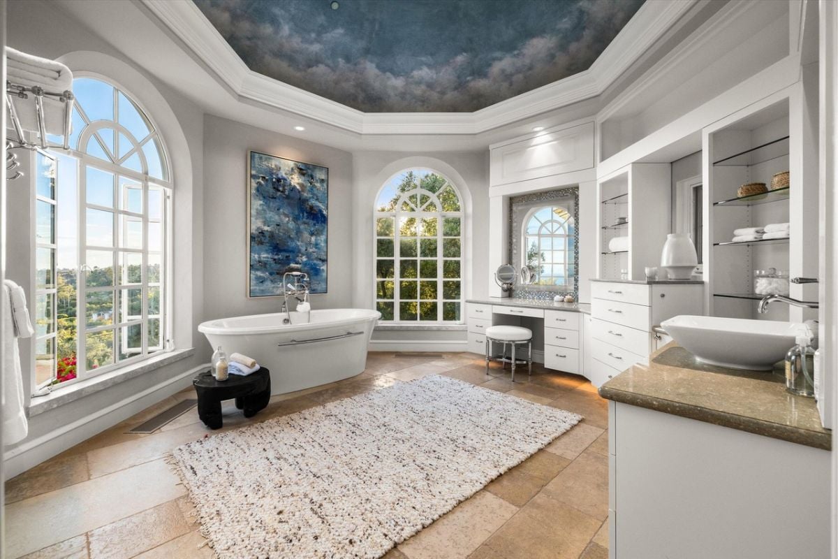 A bathroom with a painted ceiling, windows, bathtub, vanity, white cabinets, and shelves.