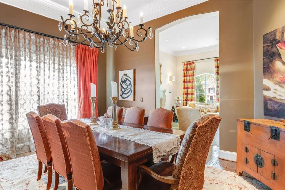 A dining room with a window with curtains, a chandelier, and a dining table surrounded by chairs.