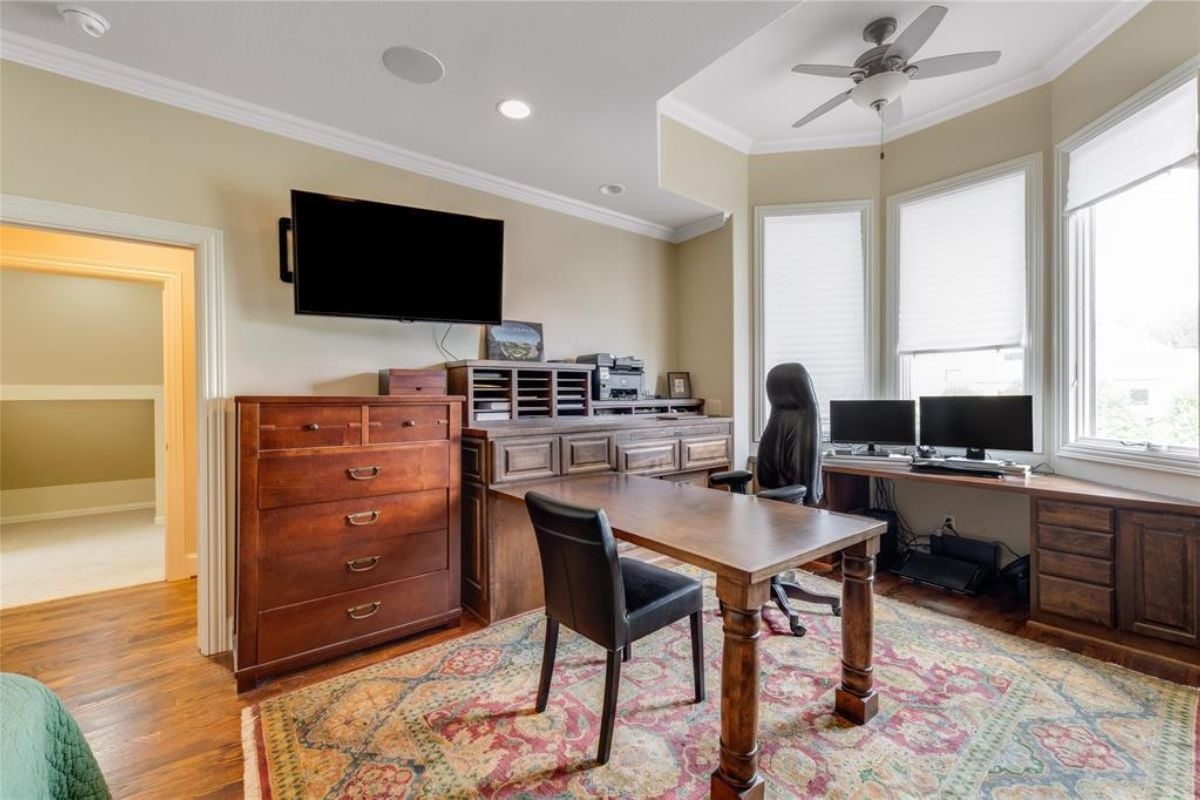 A home office with a rug, desk, chair, cabinet, windows, and a ceiling fan.