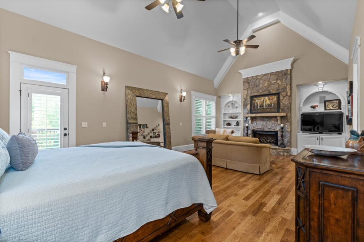 A bedroom with a bed, vaulted ceiling, ceiling fans, stone fireplace, TV, shelves, and a couch.