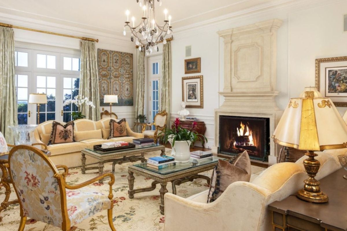 A living room with a fireplace, chandelier, window, paintings, couches, chairs, and a center table.