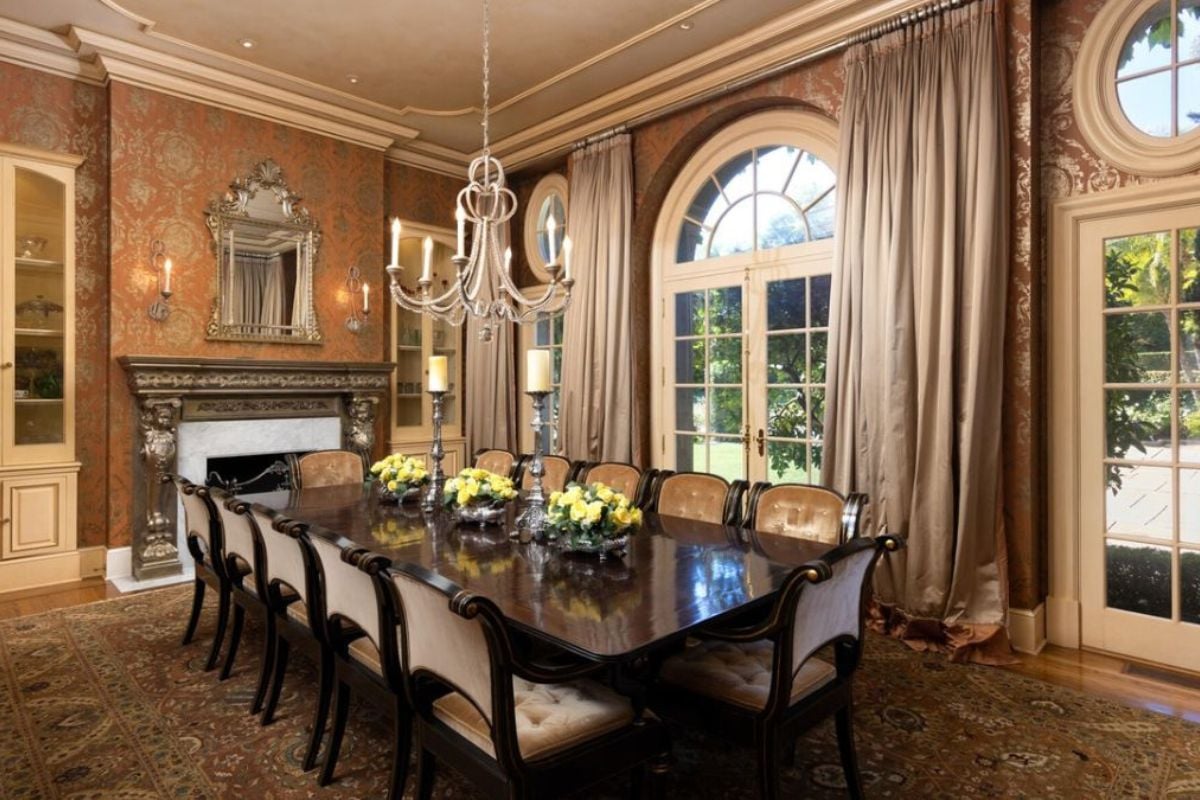 A dining room with a rug, chandelier, fireplace, windows with curtains, cabinets, and a dining table surrounded by chairs.