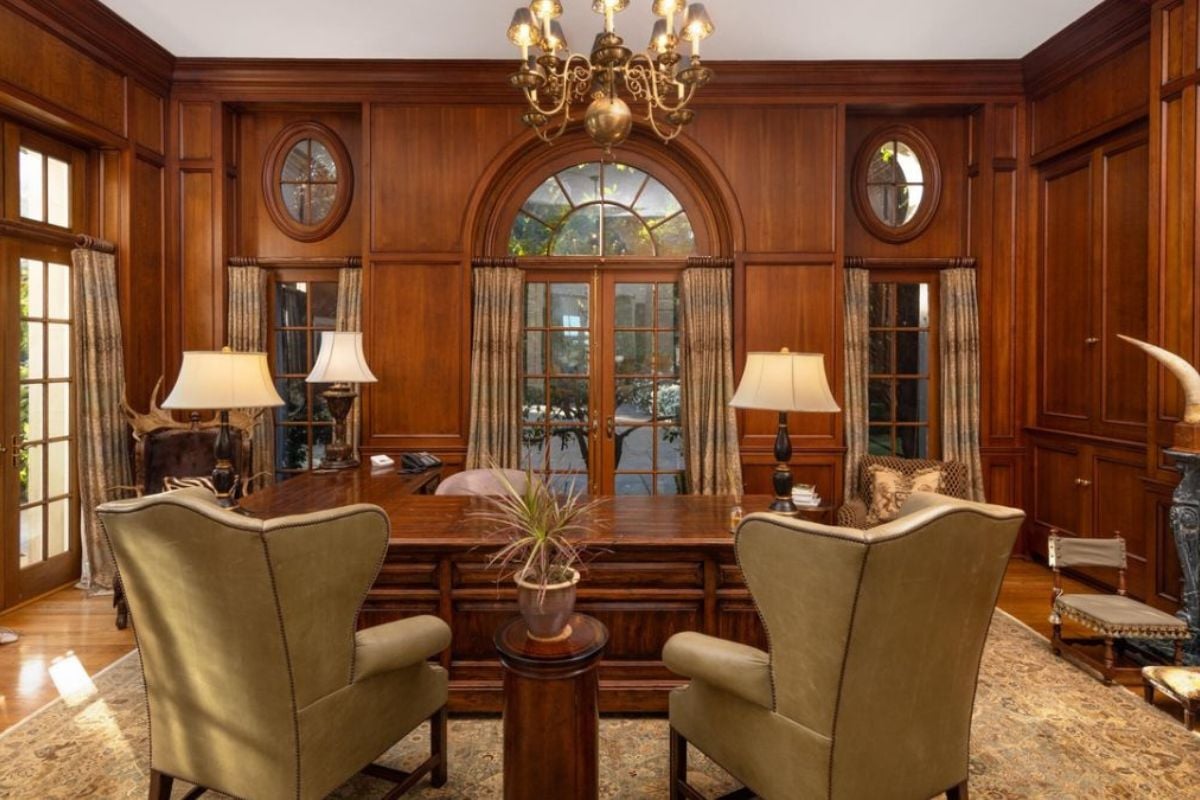 A home office with a rug, chandelier, fireplace, windows, desk, and chairs.