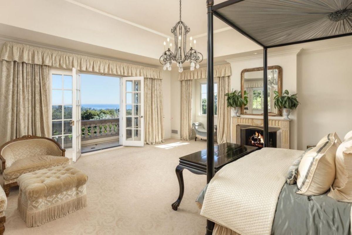 A bedroom with a canopy bed, fireplace, chandelier, mirror, chairs, and a coffee table.