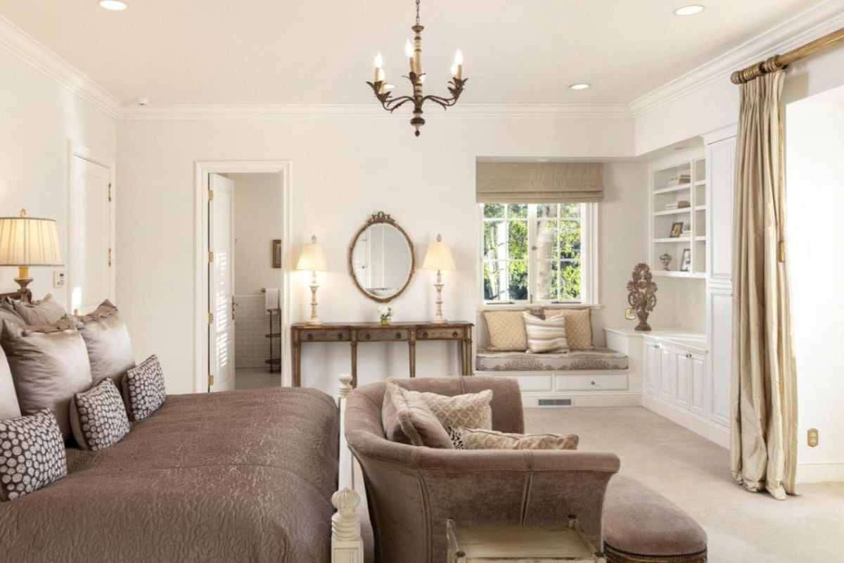 A bedroom with a bed, couch, chandelier, mirror, window, and window seating.