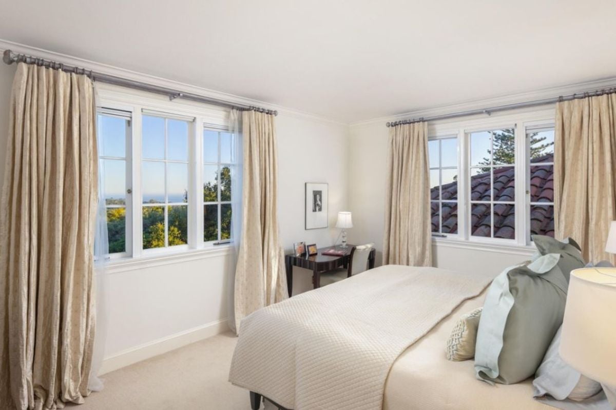 A bedroom with a bed, windows, lampshades, desk, and chair.