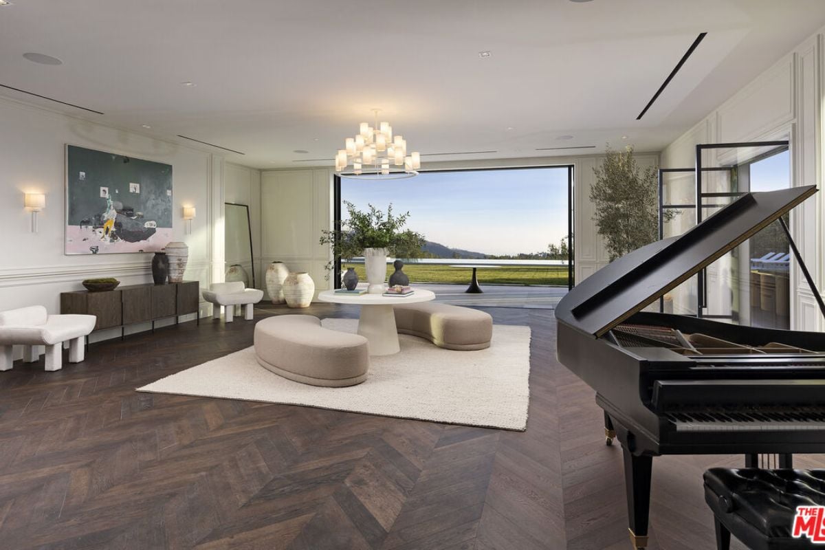 A living room with a painting, cabinet, chairs, piano, chandelier, and a center table surrounded by white curved couches.