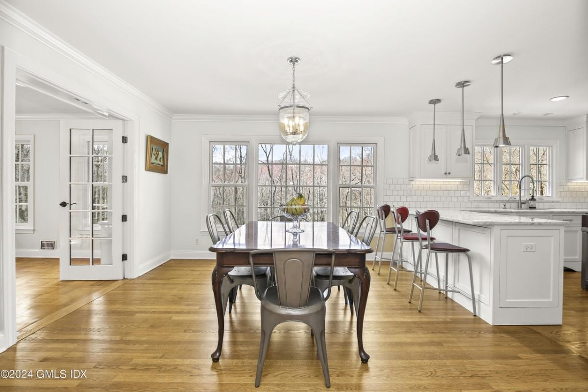 A dining area with windows, wooden floor, dining table surrounded by chairs, and kitchen.