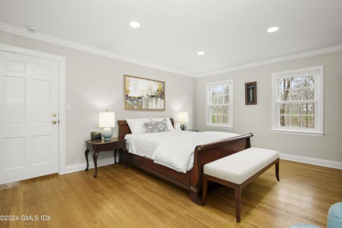 A bedroom with a bed, side table, painting, and windows.