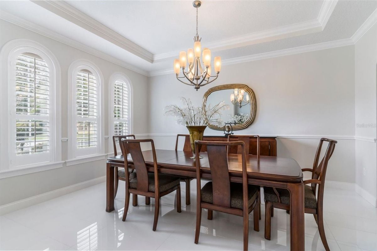 A dining room with a tiled floor, windows, chandelier, mirror, and a dining table surrounded by chairs.