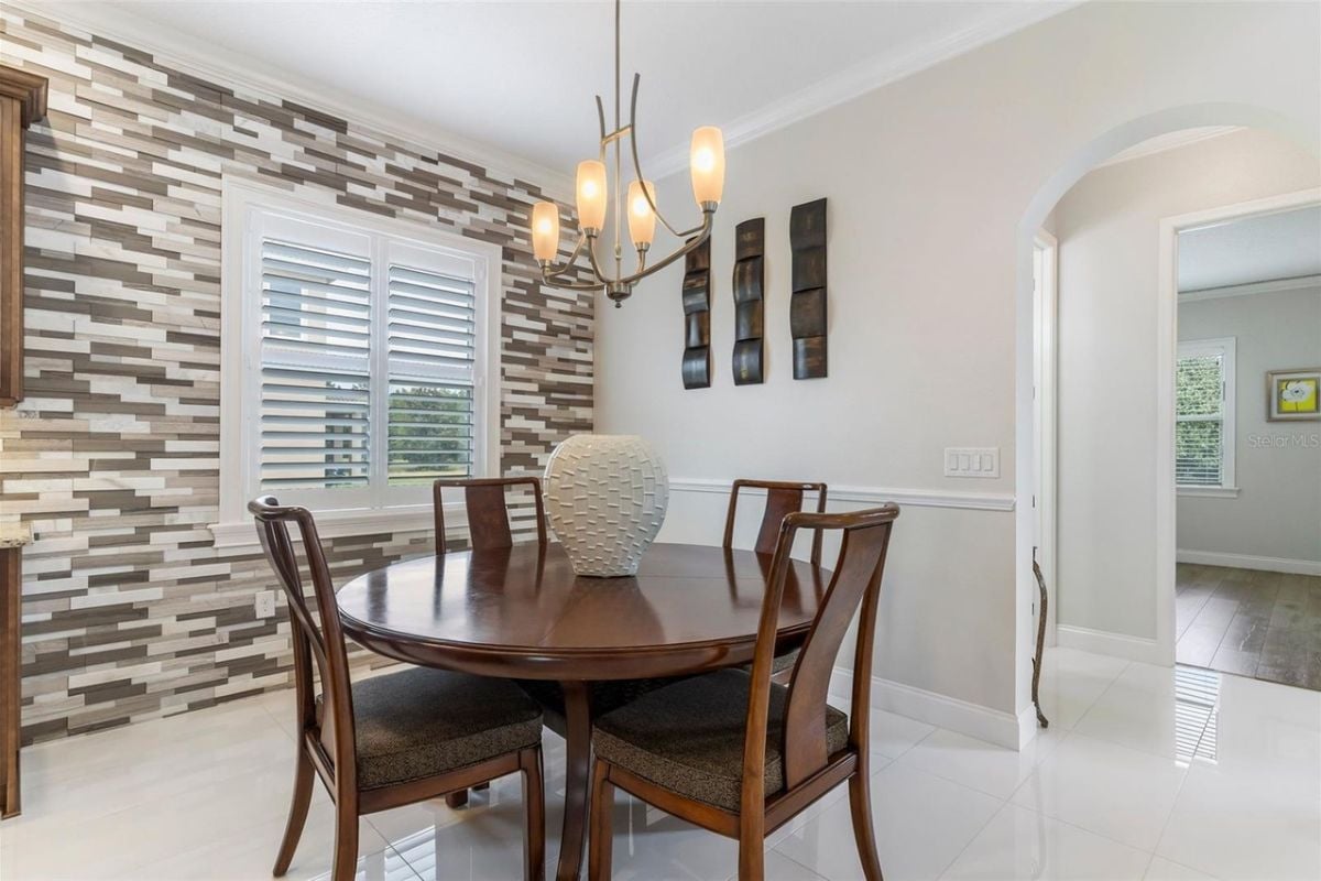 A breakfast nook with windows, a chandelier, a round table with a vase, surrounded by chairs.