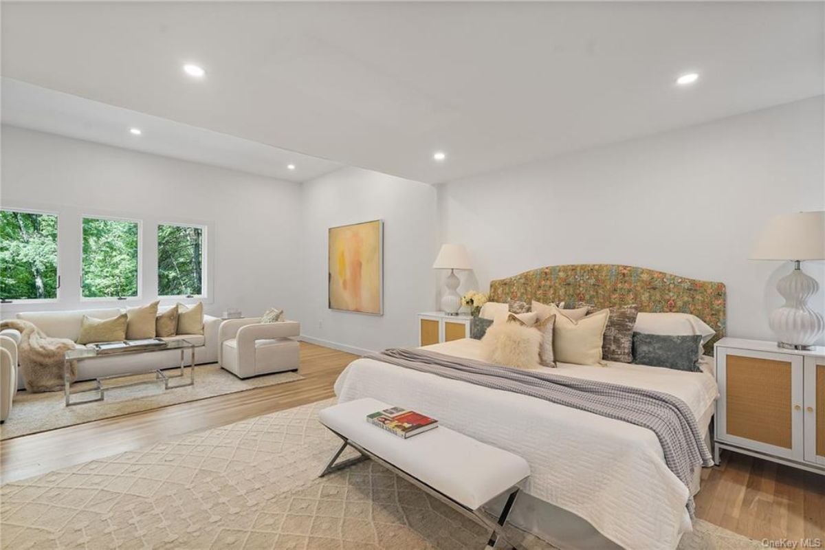 A bedroom with a bed, nightstand, windows, couch, chairs, and a glass coffee table.