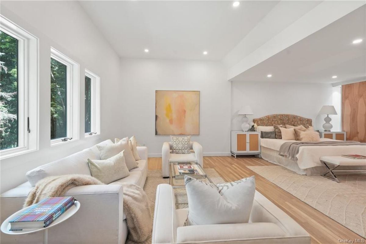 A bedroom with a bed, nightstand, windows, couch, chairs, and a glass coffee table.