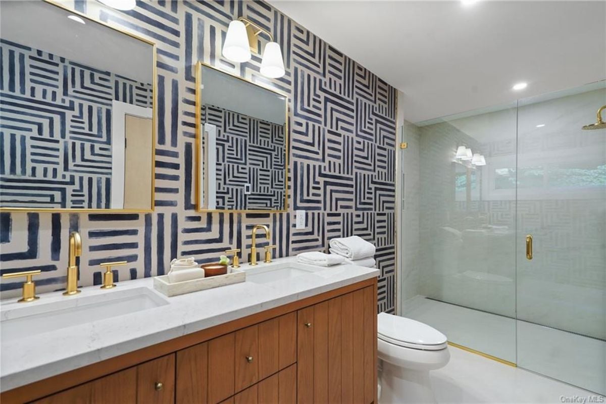 A bathroom with wallpaper, a dual vanity, a mirror, a toilet, and a shower room.