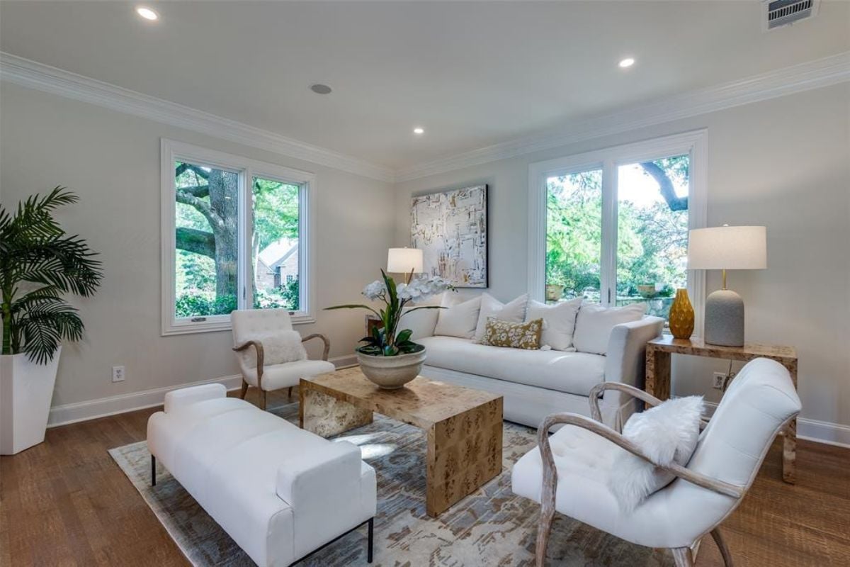 A living room with windows, houseplants, white couches, white chairs, and a center table.