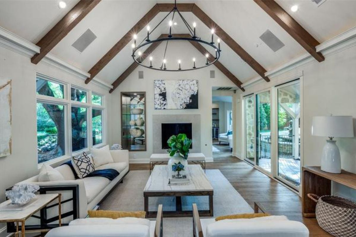 A living room with a vaulted ceiling, painting, fireplace, windows, white couch, white chairs, and a center table with a vase.