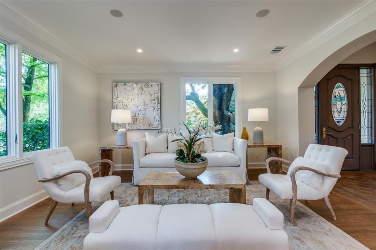 A living room with windows, a rug, white couches, white chairs, and a center table with a vase.