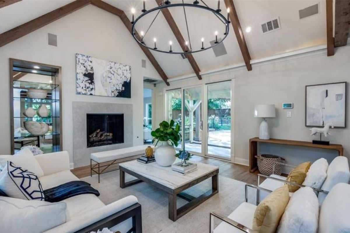 A living room with a vaulted ceiling, painting, fireplace, windows, white couch, white chairs, and a center table with a vase.